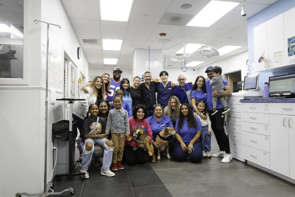 A diverse group of people, including children and adults, gathered in a veterinary clinic, smiling together. The staff, wearing blue uniforms, stands alongside pet owners and their dogs in a welcoming, modern environment.