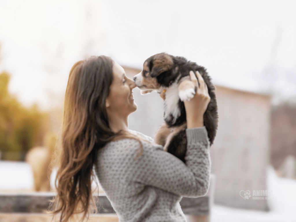 A woman joyfully holds a small puppy in her arms, sharing a tender moment outdoors. The background features a soft, blurred landscape, enhancing the warm atmosphere of their connection.