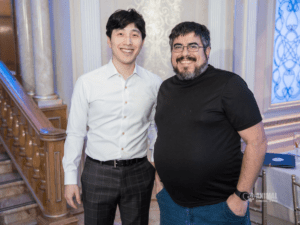 Two smiling men pose together at an event, standing near a staircase with an elegant backdrop. One man is dressed in a white dress shirt and plaid pants, while the other wears a black t-shirt with blue pants.