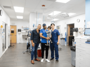 Three men stand together in a veterinary clinic, smiling. One man holds a child, while the others wear scrubs and display a friendly demeanor. The clinic features modern equipment and a clean, bright environment.