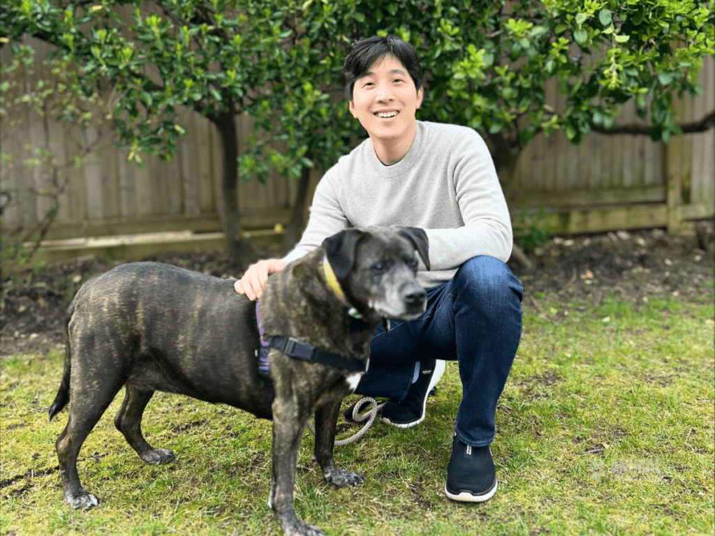 Dr. Kim in a light gray sweater crouching next to a dog in a grassy backyard surrounded by greenery.